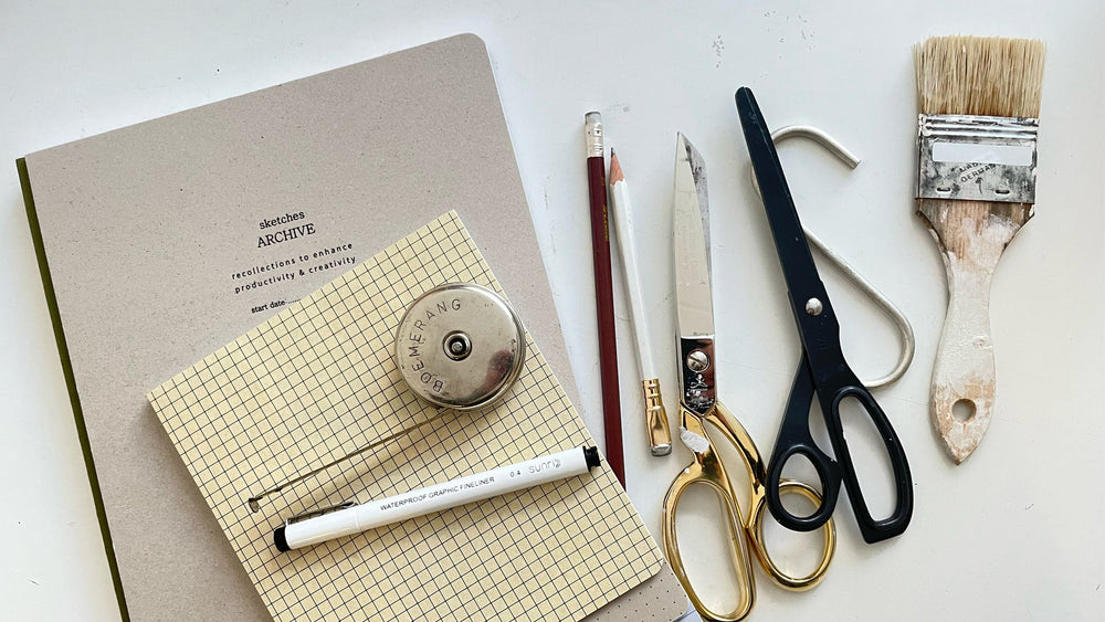 flat lay of sketchbook and grid notebook with fineliner pen, pencil, metal tape measure, two scissors and a paintbrush on a table