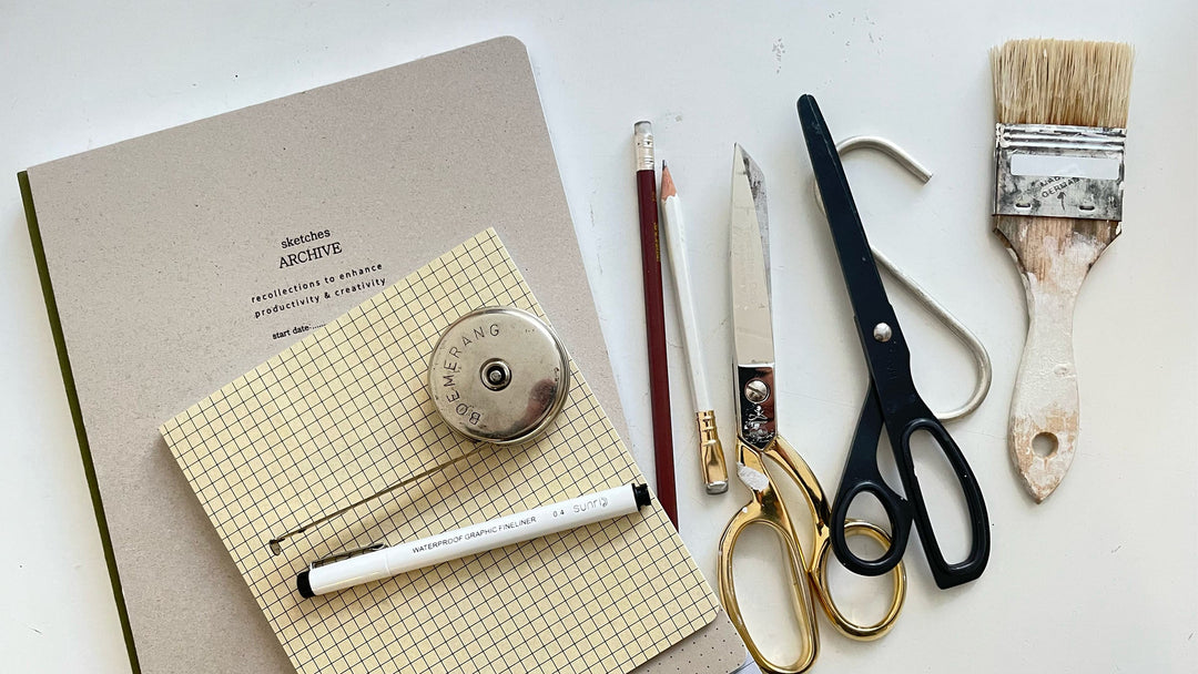 flat lay of sketchbook and grid notebook with fineliner pen, pencil, metal tape measure, two scissors and a paintbrush on a table