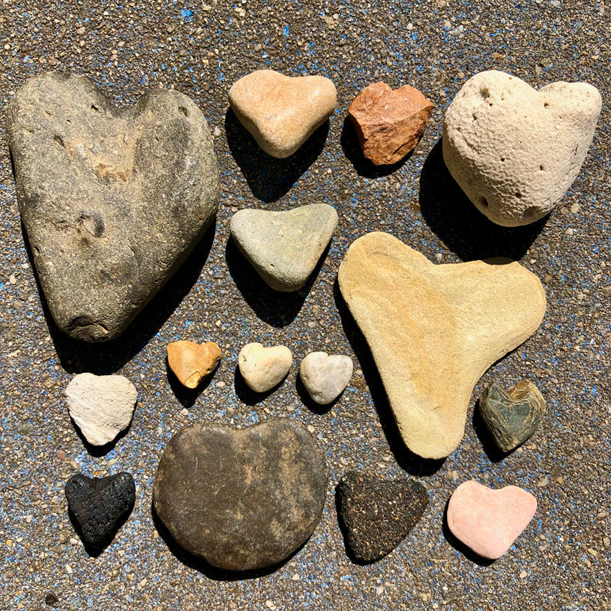 A collection of heart-shaped and irregularly shaped smooth river stones in various colors, including beige, gray, brown, pink, black, and white, arranged on a textured gray asphalt surface.