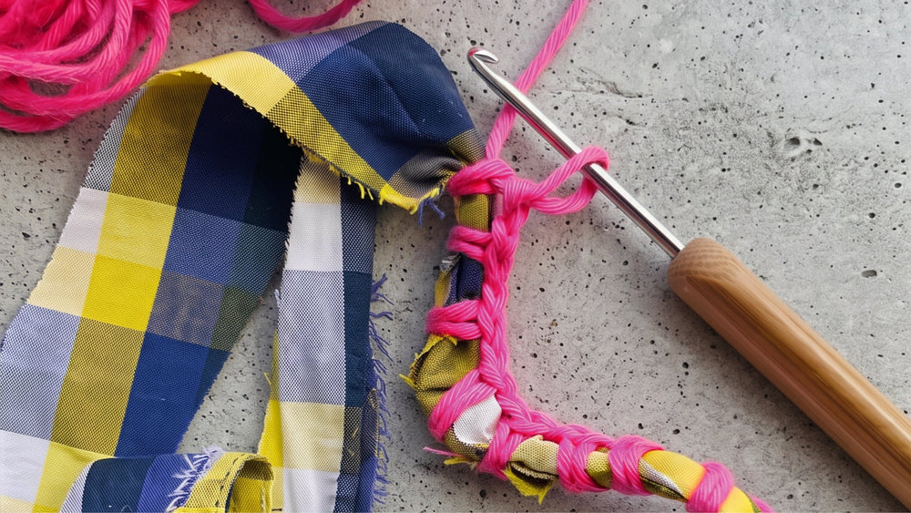 A close-up of a crochet hook with a wooden handle working through bright pink yarn, with a plaid fabric strip in navy, yellow, white, and gray beside it on a concrete surface.