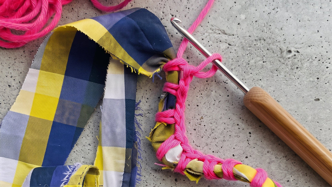 A close-up of a crochet hook with a wooden handle working through bright pink yarn, with a plaid fabric strip in navy, yellow, white, and gray beside it on a concrete surface.