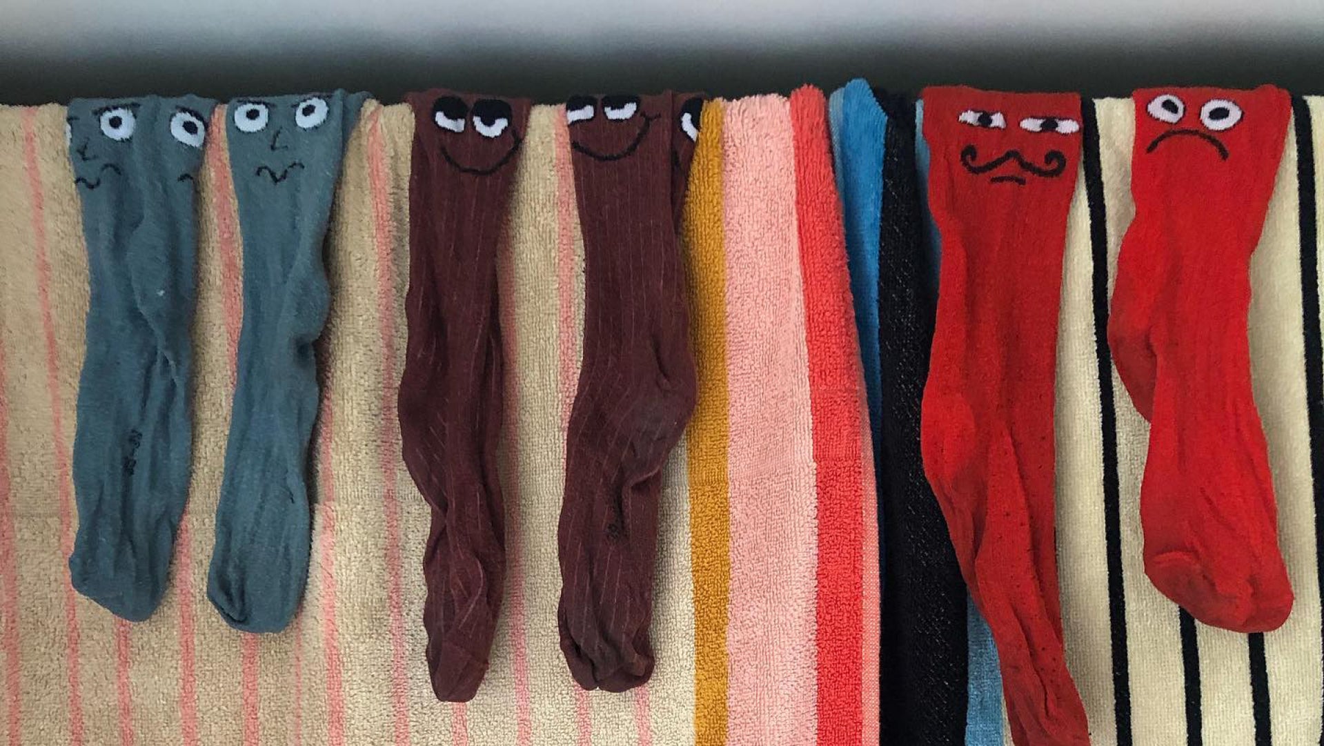 Six novelty socks with hand-painted cartoon faces, arranged vertically on a striped towel. The socks come in muted blue-gray, dark brown, peach, black, and bright red, with some featuring mustaches or winking eyes.