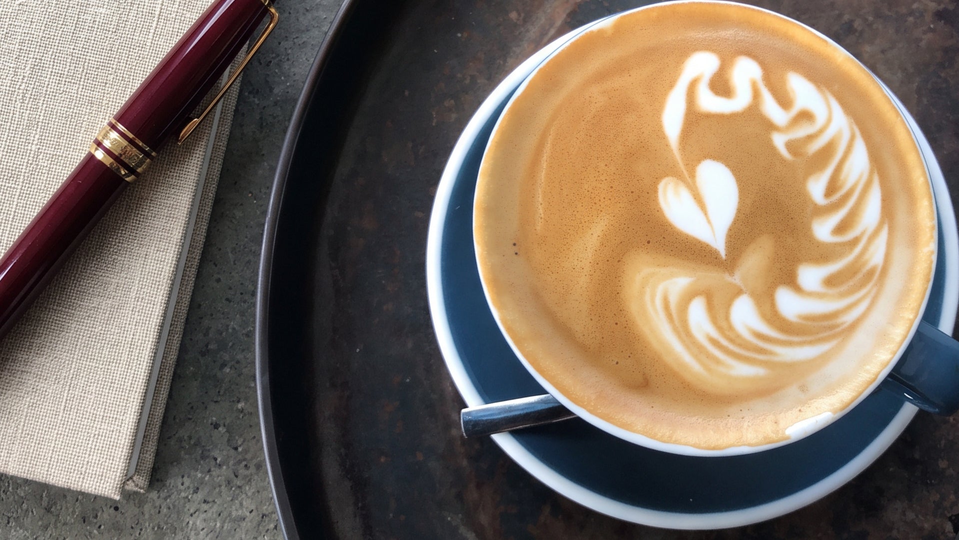 A top-down view of a cup of latte art coffee on a dark tray, next to a notebook and a dark red pen.