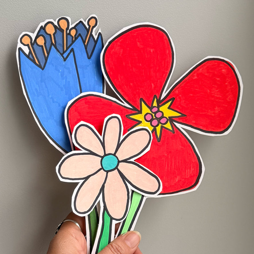 A hand holding three hand-drawn paper flowers in red, blue, and light pink with black outlines, set against a plain gray wall.