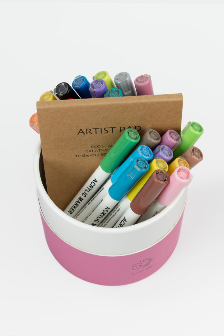 Open recycled cardboard pink tube showing assorted acrylic markers and an artist pad
