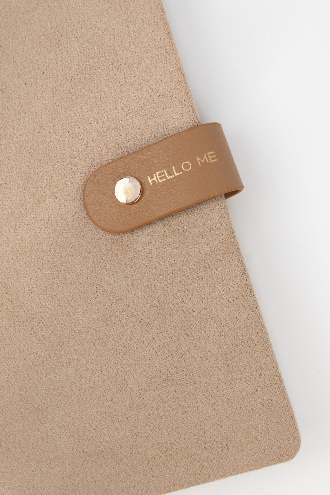 detail of brown a faux-suede notebook with a rounded flap closure with gold button detail and hello me text