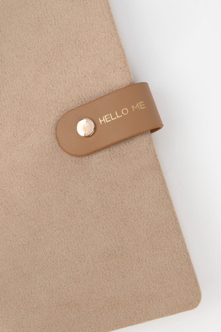 detail of brown a faux-suede notebook with a rounded flap closure with gold button detail and hello me text