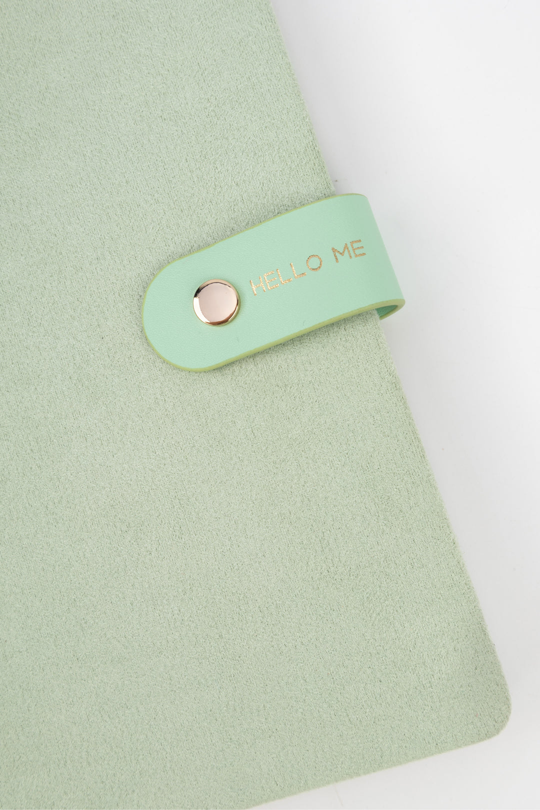 detail of a mint faux-suede notebook with a rounded flap closure with gold button detail and hello me text