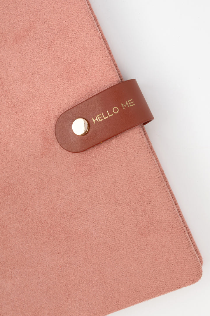 detail of a pink faux-suede notebook with a rounded flap closure with gold button detail and hello me text