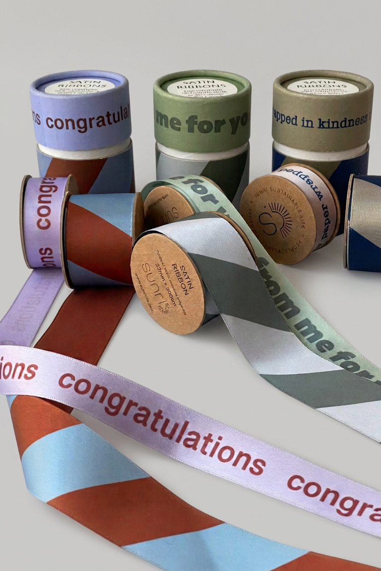 A group of decorative ribbon spools and loose rolls with printed congratulatory phrases, arranged on a light gray background. The ribbons feature geometric color-blocked patterns in purple, green, brown, blue, and white, with some spools showing kraft paper cores.