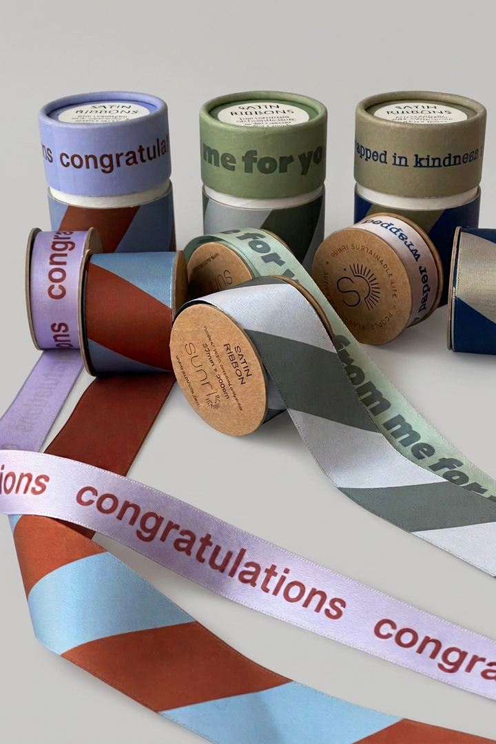 A group of decorative ribbon spools and loose rolls with printed congratulatory phrases, arranged on a light gray background. The ribbons feature geometric color-blocked patterns in purple, green, brown, blue, and white, with some spools showing kraft paper cores.