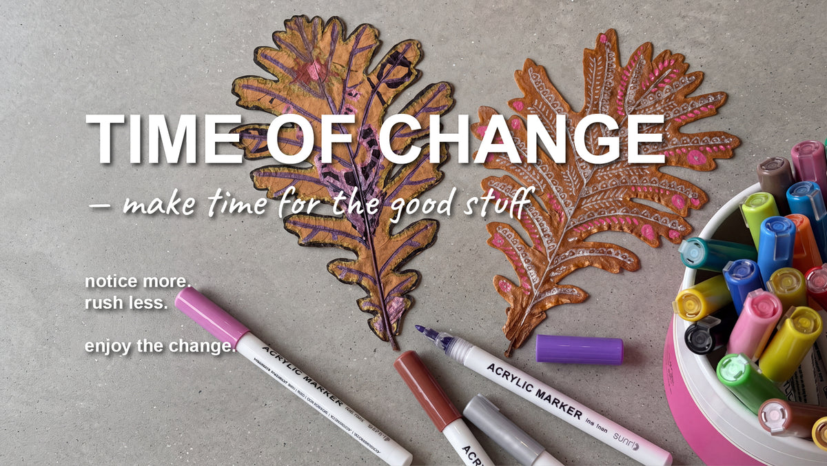 A styled flat-lay featuring two hand-painted oak leaves with gold glitter accents, surrounded by acrylic markers on a textured gray surface. Large white text reads 'TIME OF CHANGE' with the subtitle '- make time for the good stuff'.