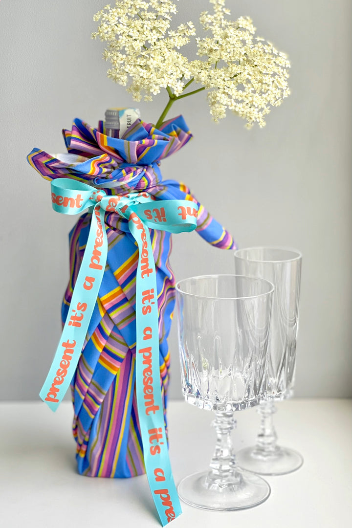 A wrapped gift box tied with a bright blue ribbon printed with the words 'present it's a present' in orange, styled with white baby's breath flowers and two clear glass goblets on a white surface against a light gray background.