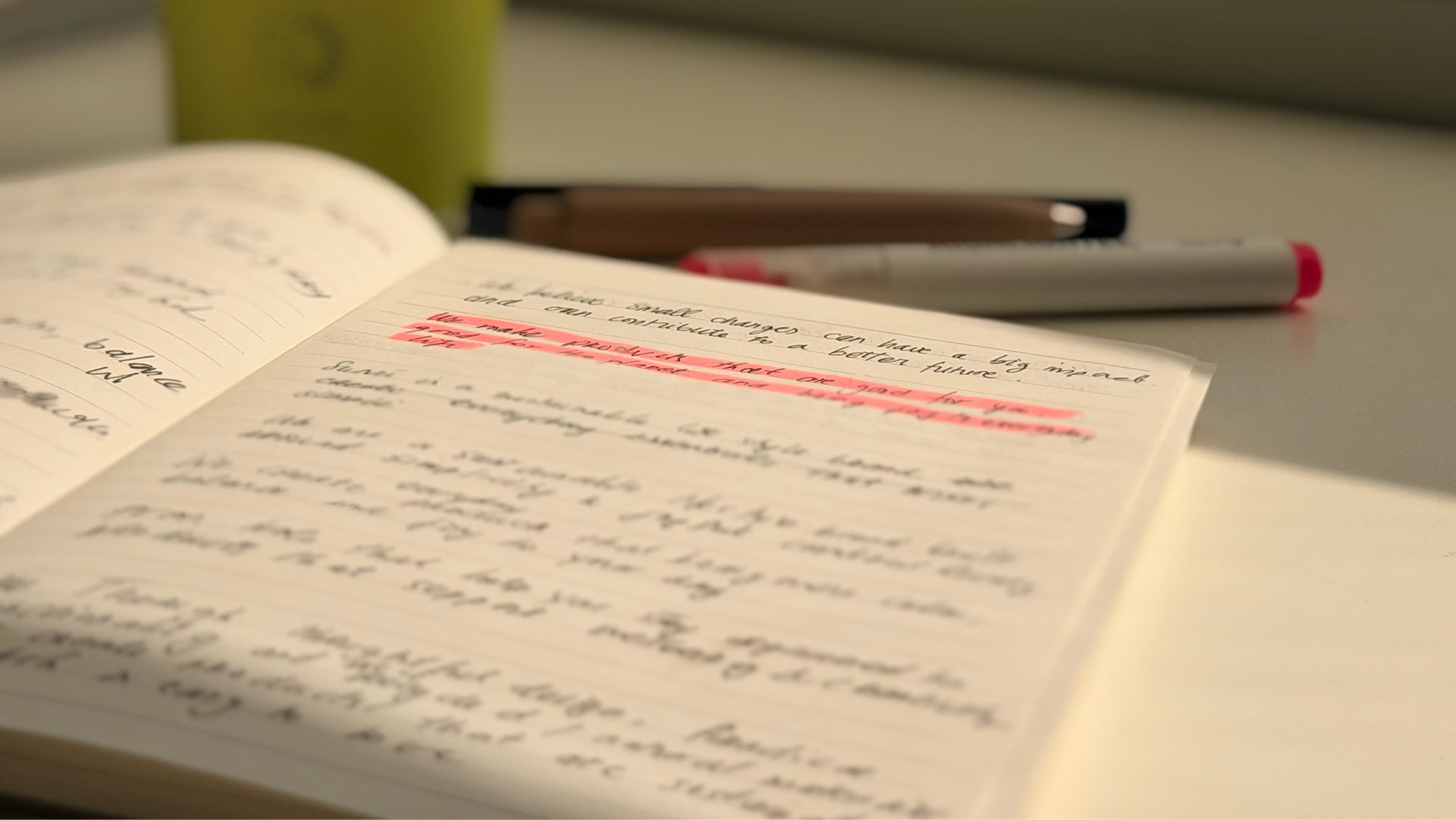 An open handwritten notebook with cursive writing and pink highlighter marks on the page, with two pens resting on the desk in the background.