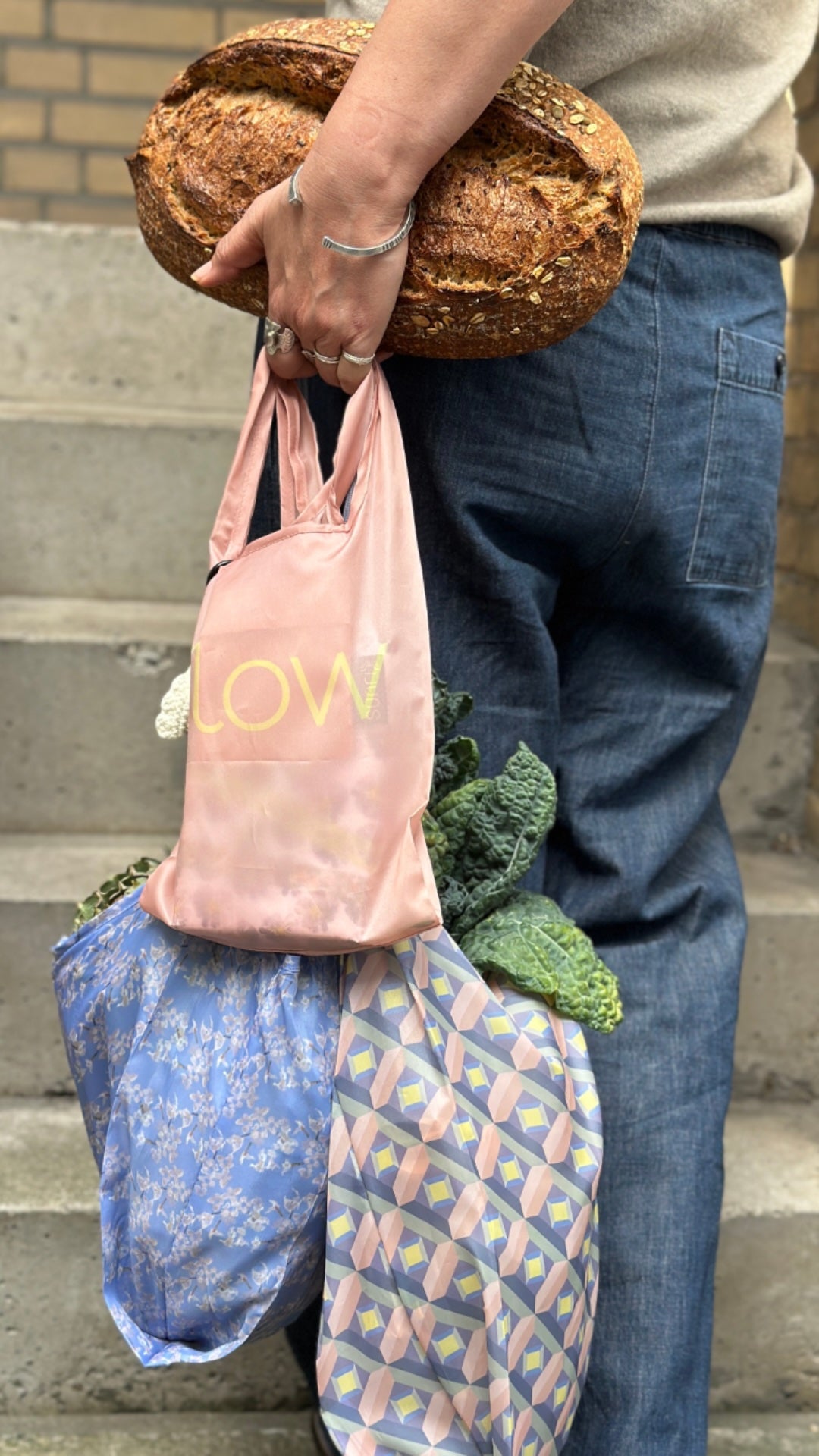 A person carrying a loaf of bread and multiple reusable shopping bags on their arm while standing on concrete stairs. The bags include a dusty pink bag with yellow text, a blue floral bag, and a pink and blue geometric patterned bag.