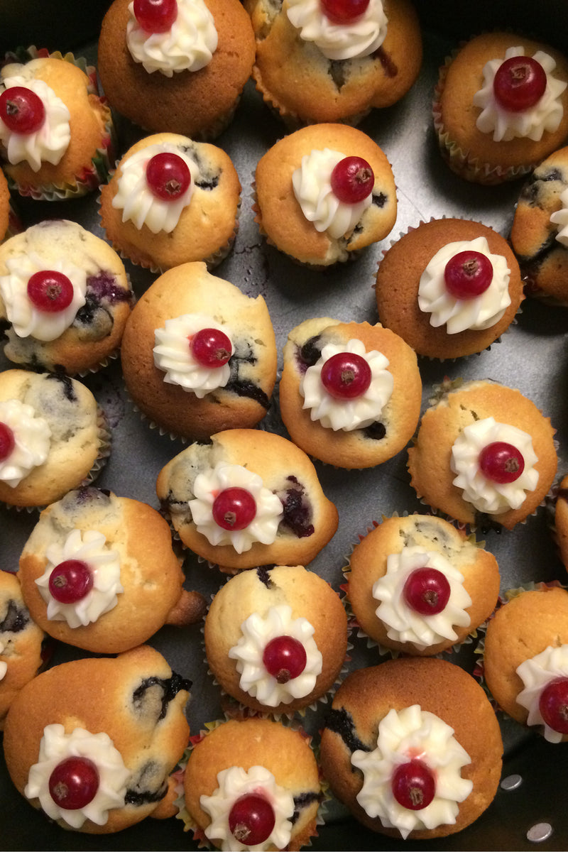A close-up overhead view of multiple small blueberry muffins topped with white whipped frosting and a single red berry, arranged closely together on a dark tray.