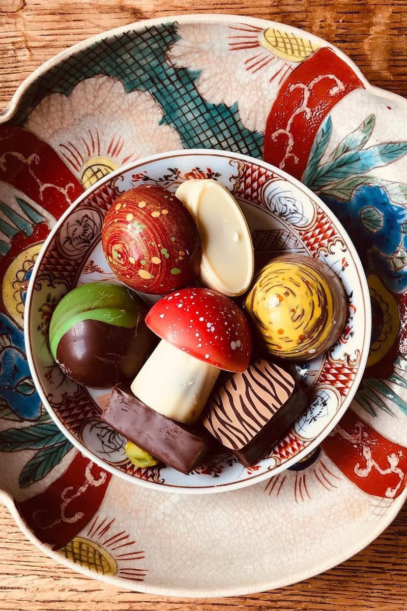A bowl of assorted gourmet chocolates and confections arranged on a decorative ceramic plate, set on a wooden surface. The chocolates include mushroom-shaped pieces, striped truffles, and patterned dark and milk chocolate pieces.