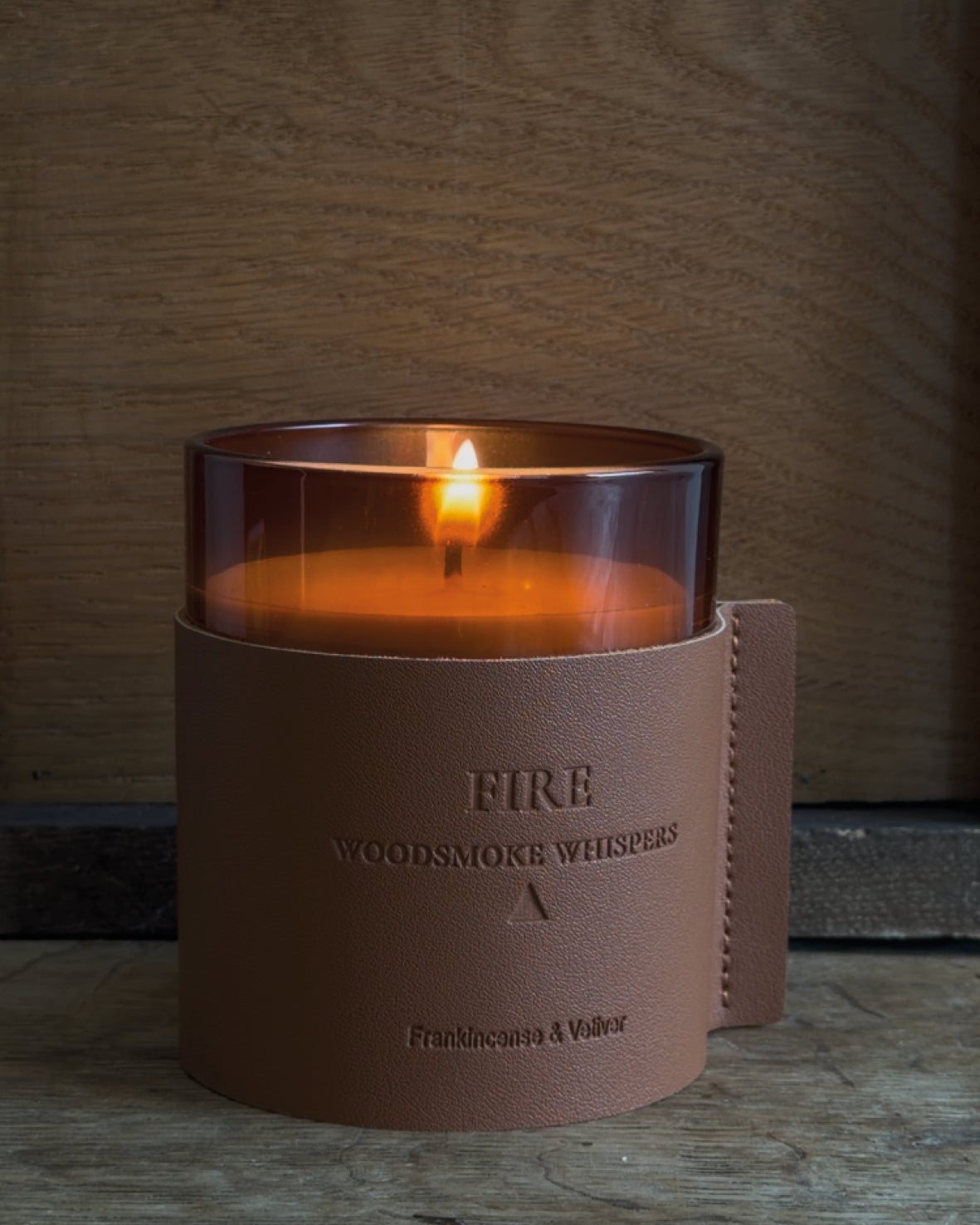 A amber glass jar candle with white wax sits in a brownish faux leather sleeve with a natural background