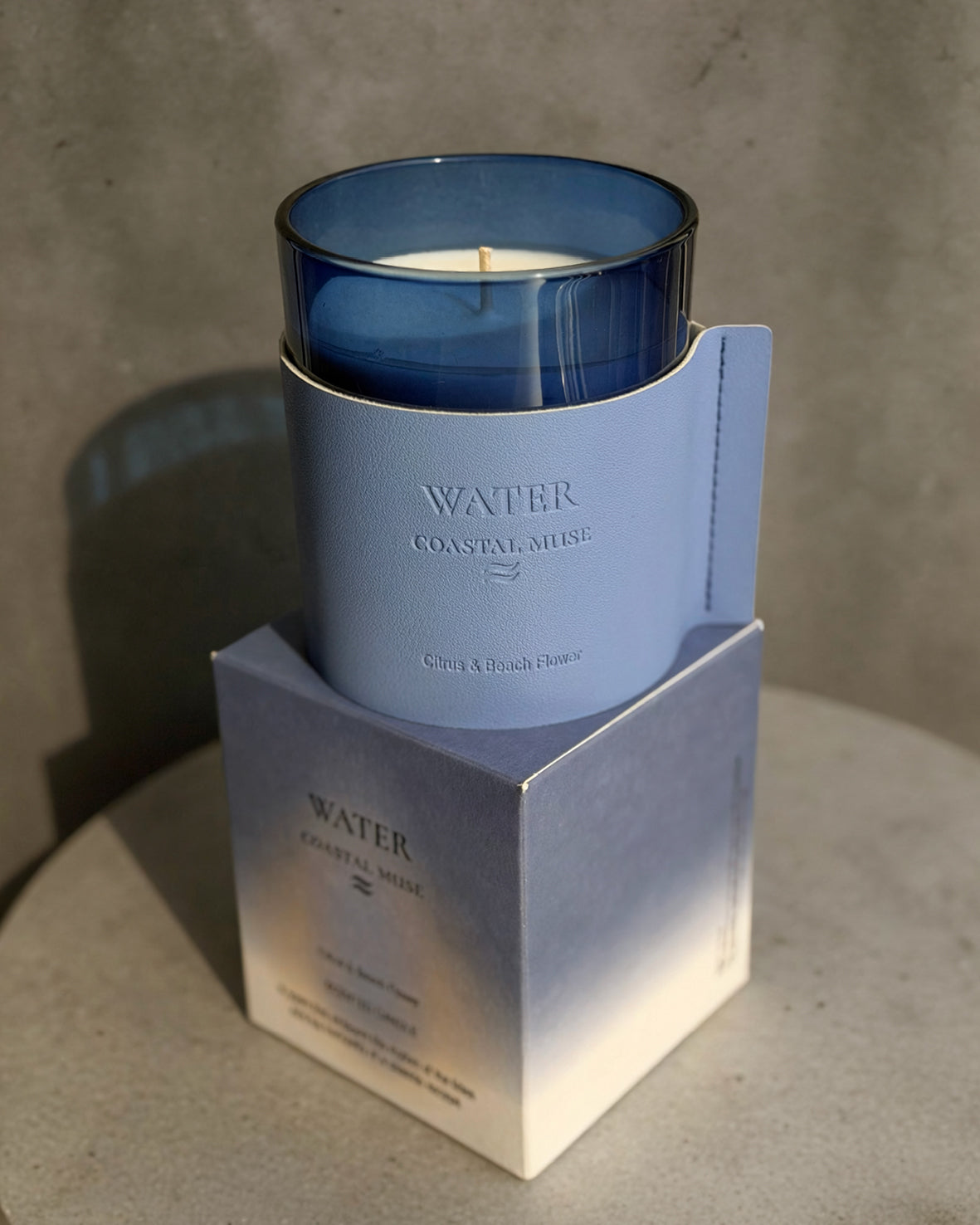 A blue glass jar candle with white wax sits in a light blue paper sleeve, displayed on top of its matching blue and white box against a textured gray background.