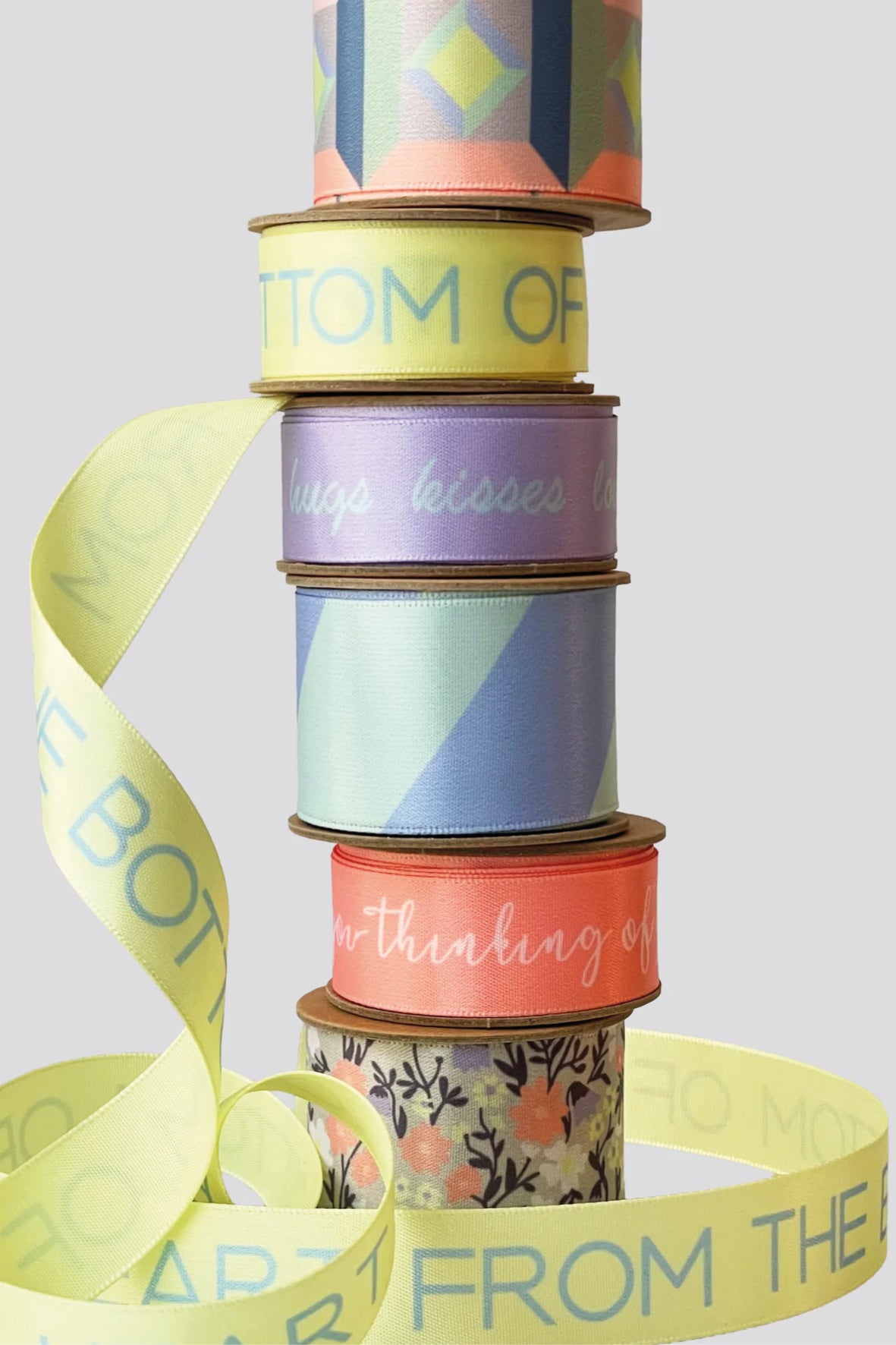 A stack of colorful patterned and printed ribbons wound vertically, with loose ribbons draped around the base on a light gray background.