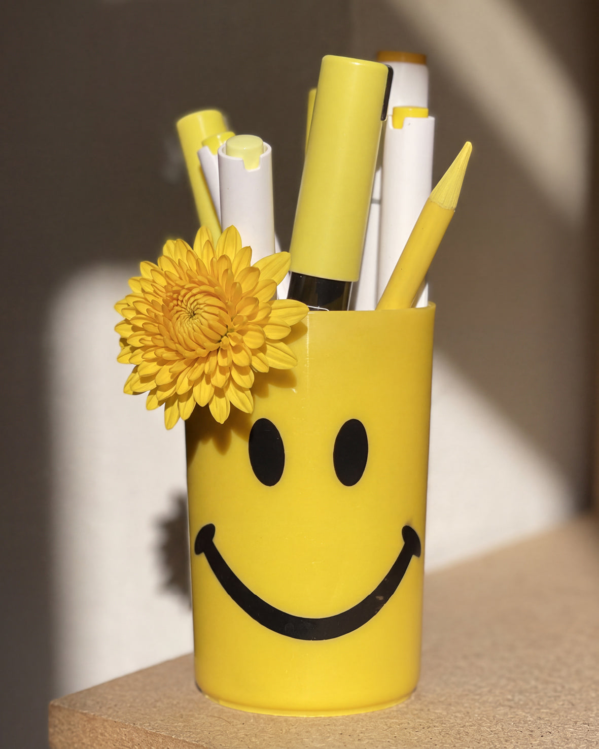 A yellow smiley face pencil holder cup filled with pens and a yellow flower, sitting on a wooden surface with warm sunlight and shadows.