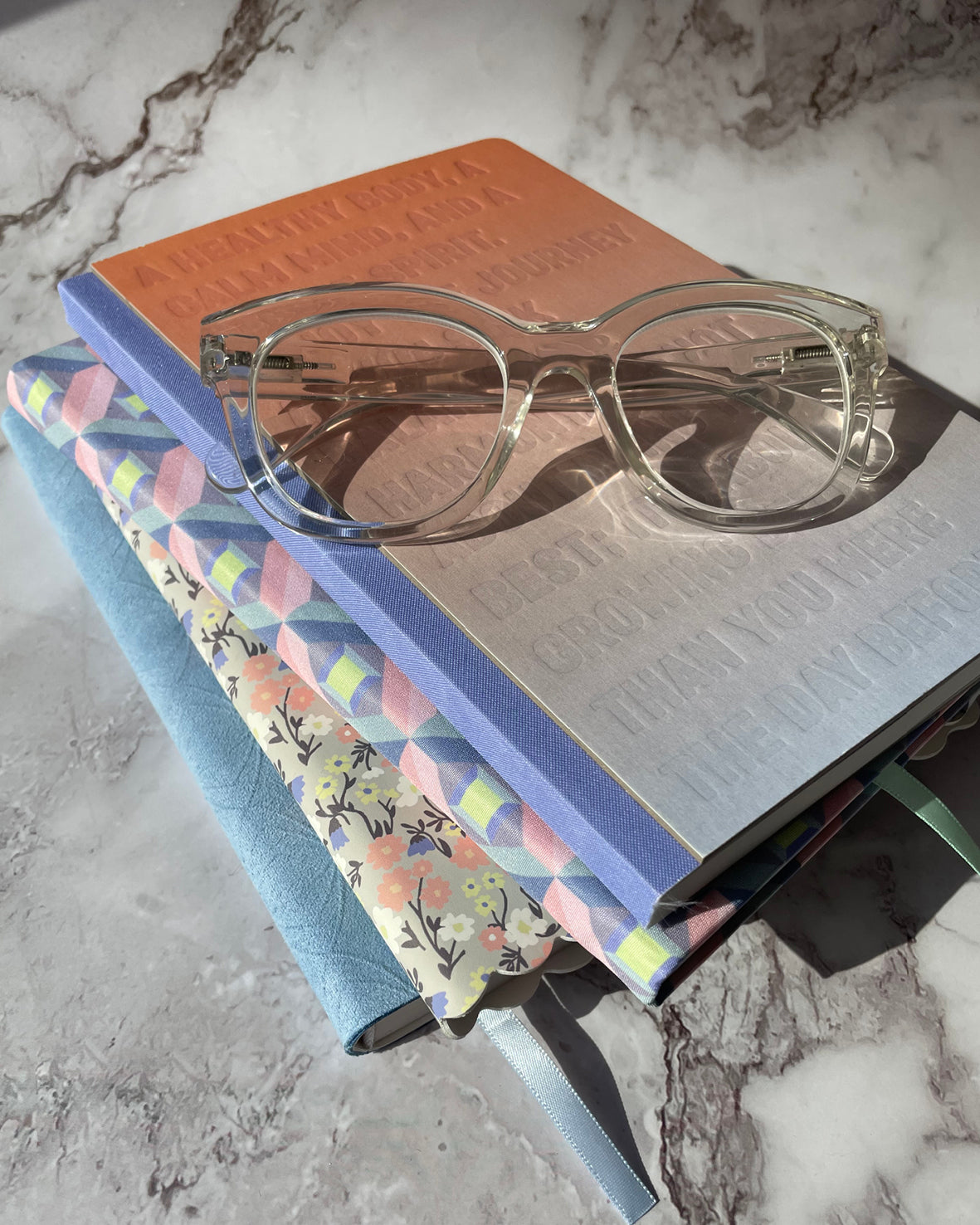 Clear eyeglasses resting on a stack of notebooks on a gray and white marble surface. The notebooks include a peach-colored one, a blue one, and a floral-patterned pastel one.