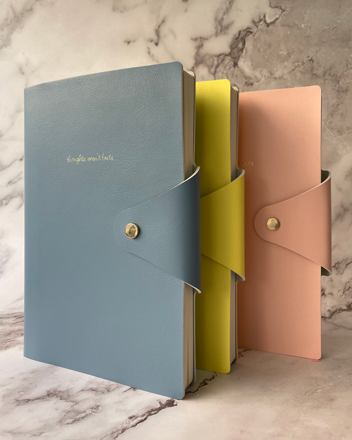 Three slim leather notebooks or journals standing upright on a marble surface: a light blue one in front with gold text reading 'beautiful book,' a bright yellow one behind it, and a soft peach-pink one behind that. Each has a flap closure with a metal snap and a matching side strap.