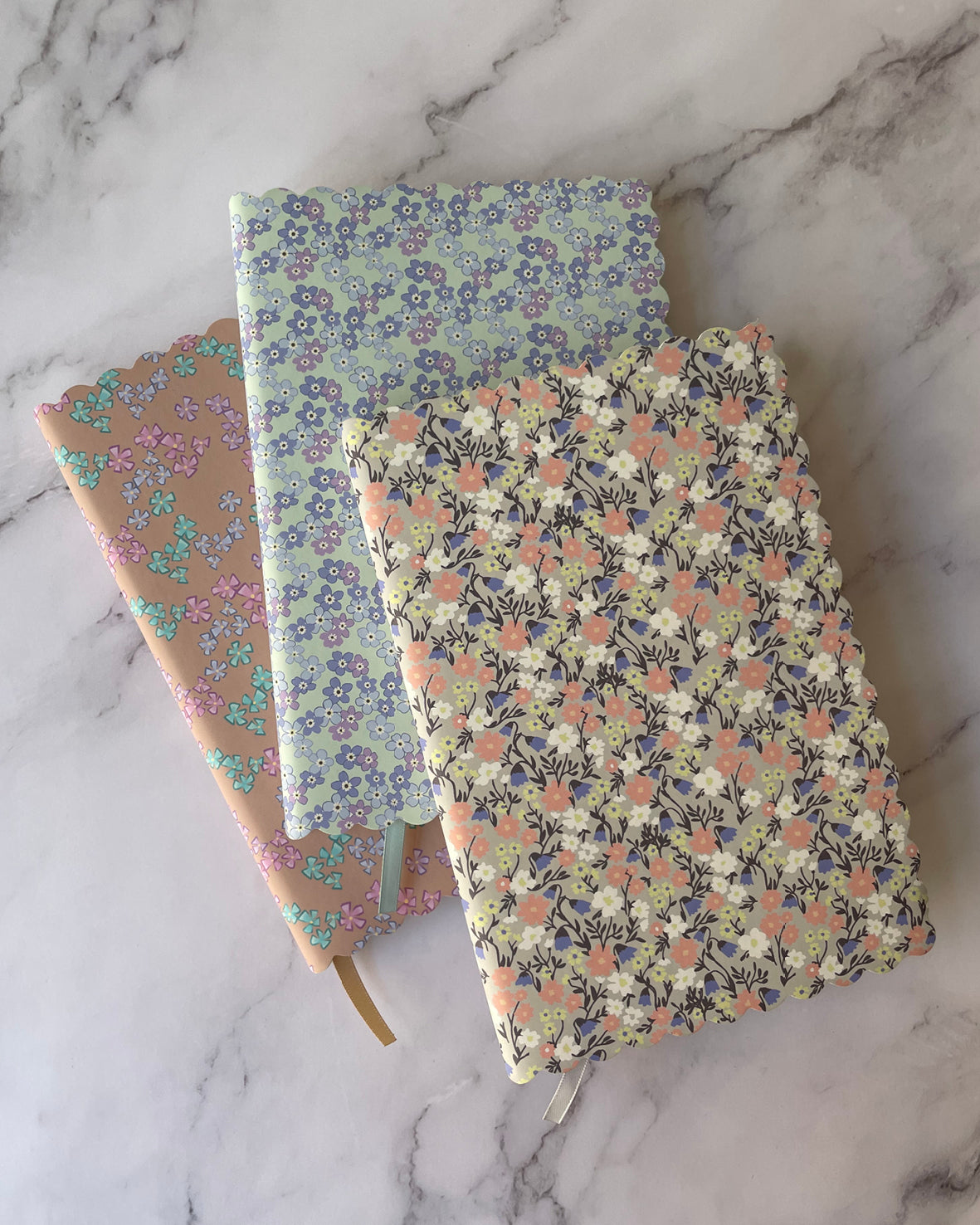 Three floral patterned notebooks stacked on a white marble surface. The top notebook has a dense multicolor floral print, the middle notebook has a light blue and purple floral pattern, and the bottom notebook has a peach and pink floral pattern with a gold ribbon bookmark.