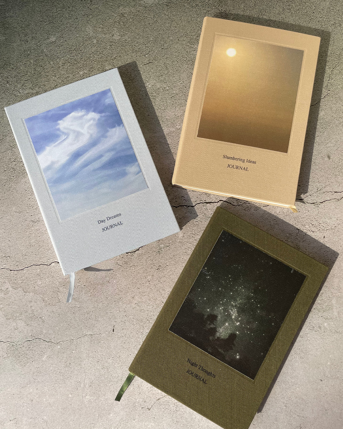 Three hardcover journals with Polaroid-style photo windows are arranged on a textured concrete surface. One journal is white with a blue sky cover, one is beige with a golden sunset cover, and one is olive green with a starry night sky cover.
