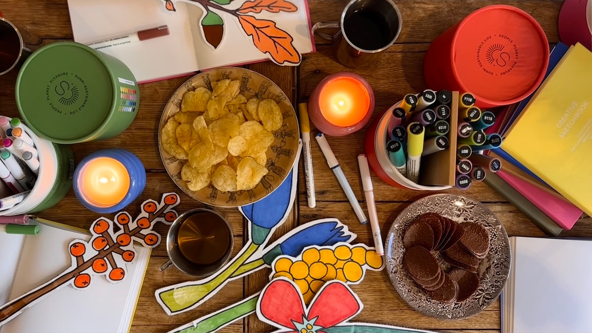 A styled flat-lay of multiple colorful circular tins, likely home fragrance or gift containers, arranged on a wooden table alongside candles, snacks, and stationery.