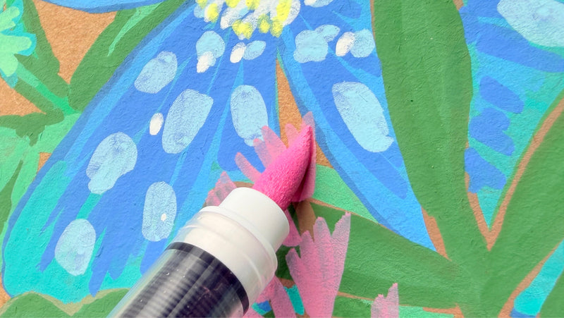 A close-up of a paint marker with a pink sponge tip being used to paint a blue flower with light blue spots on a colorful painted surface.