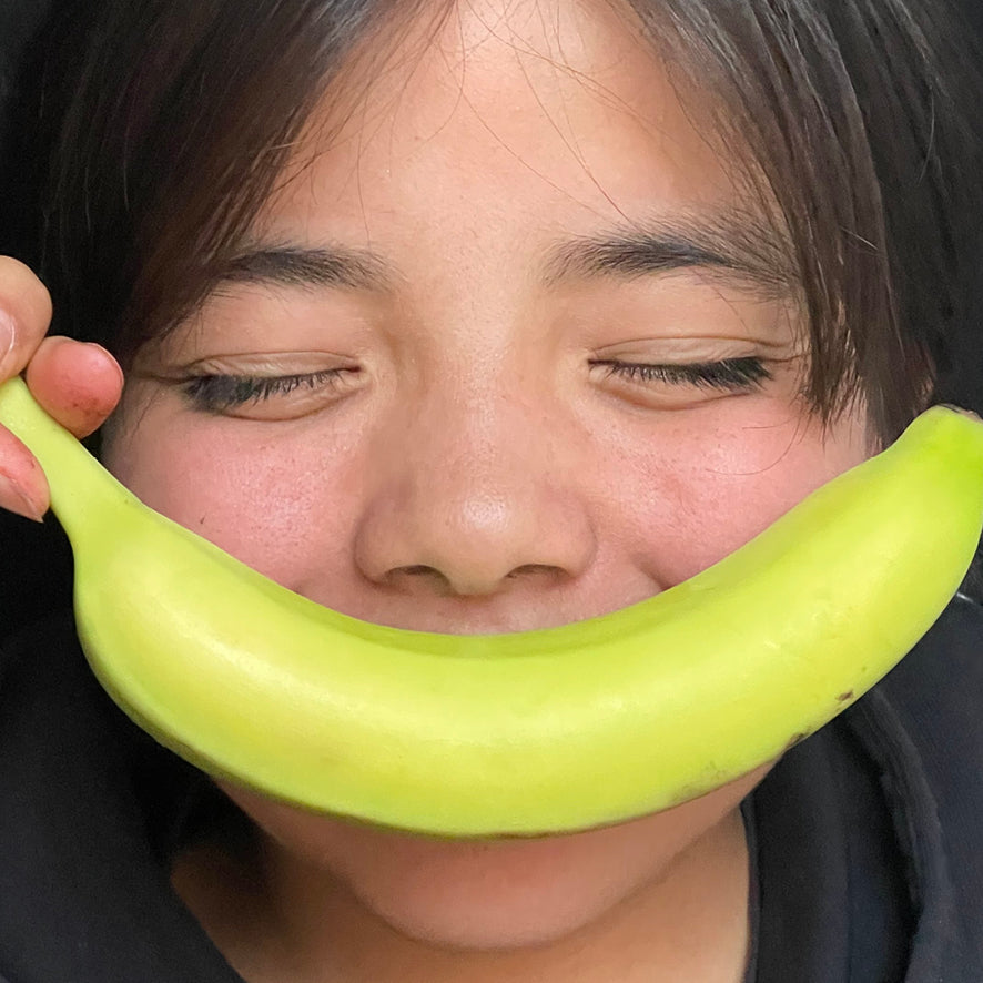 A close-up of a person holding a bright green banana in front of their mouth, with their eyes closed and a playful expression.