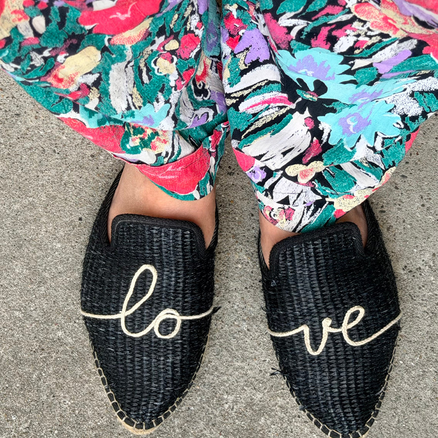 A top-down view of a person wearing black woven espadrille-style slip-on shoes with the word "love" embroidered in white script across the fronts, paired with colorful floral pants on a gray pavement background.
