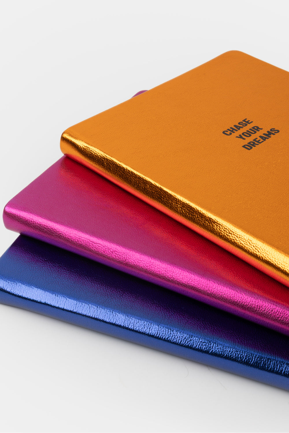 Three metallic notebooks stacked in orange pink and blue with the orange notebook on top
