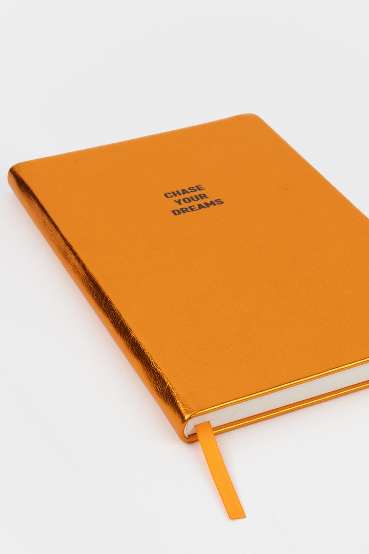 Angled view of closed orange metallic A5 notebook with chase your dreams text
