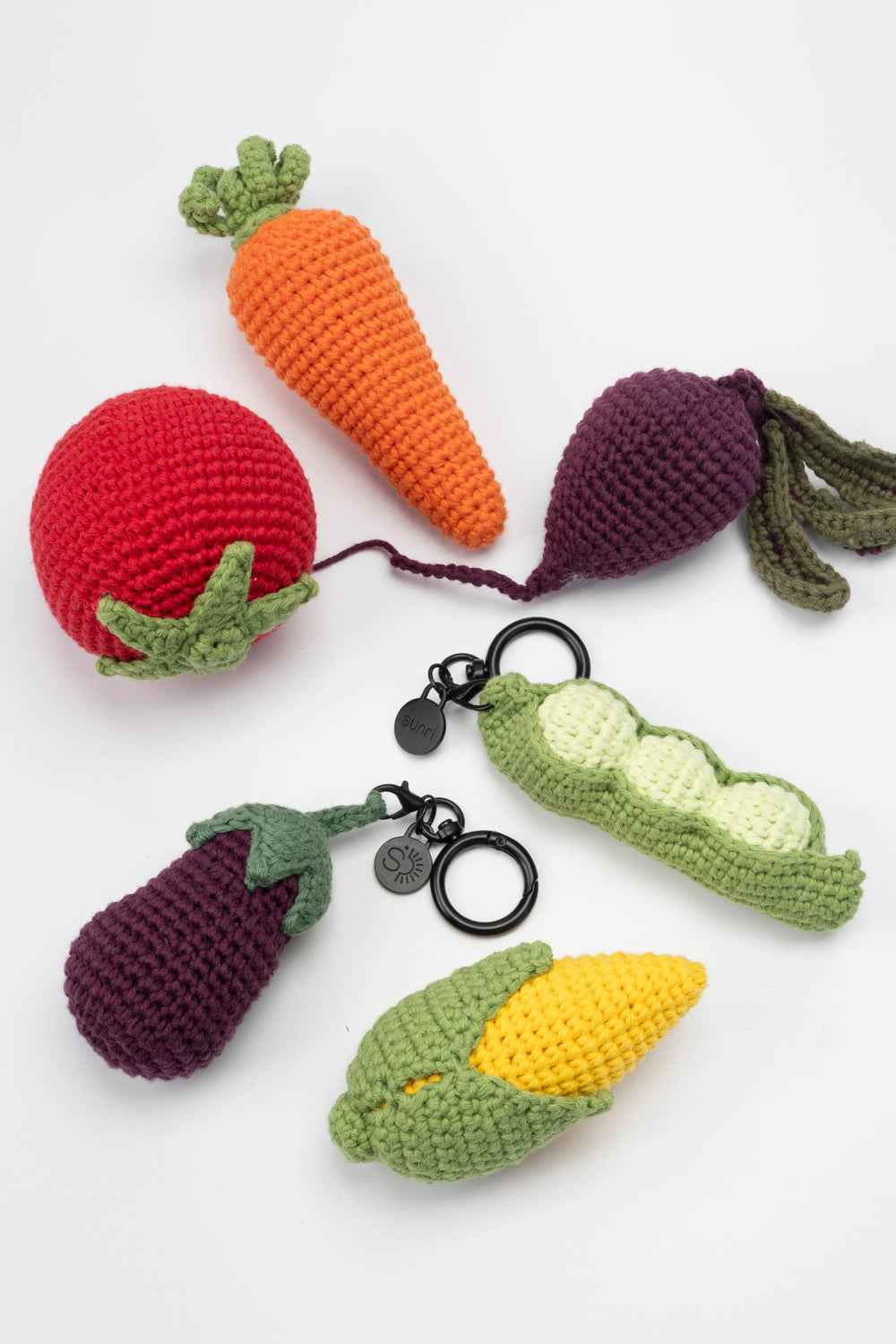Set of crocheted vegetable keychains including a carrot, radish, pea pod, eggplant, and corn on a white background.