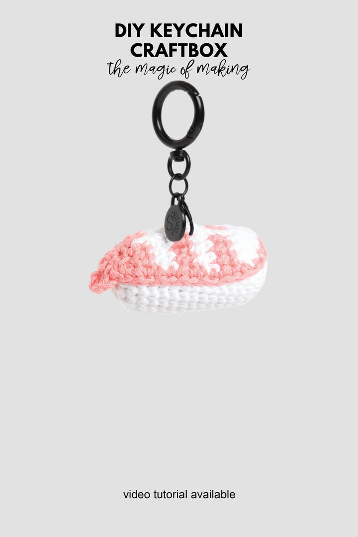 Crocheted keychain with pink and white design on a gray background, featuring text about DIY keychain craftbox.