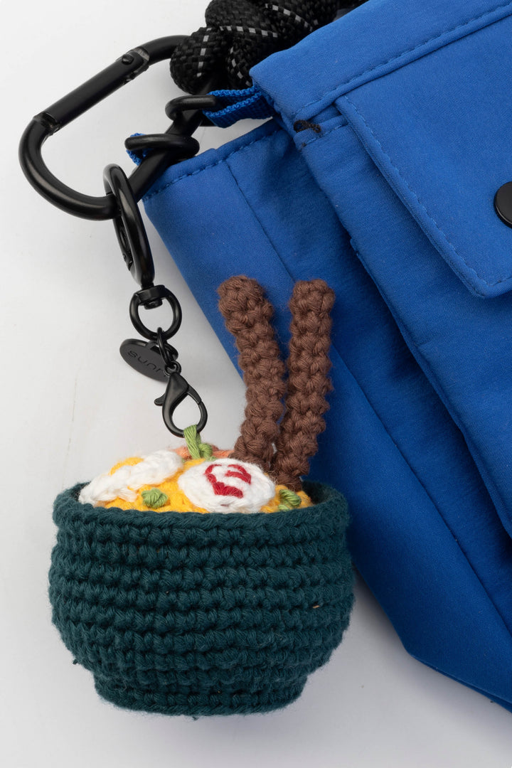 Crocheted ramen keychain attached to a blue bag on a white background