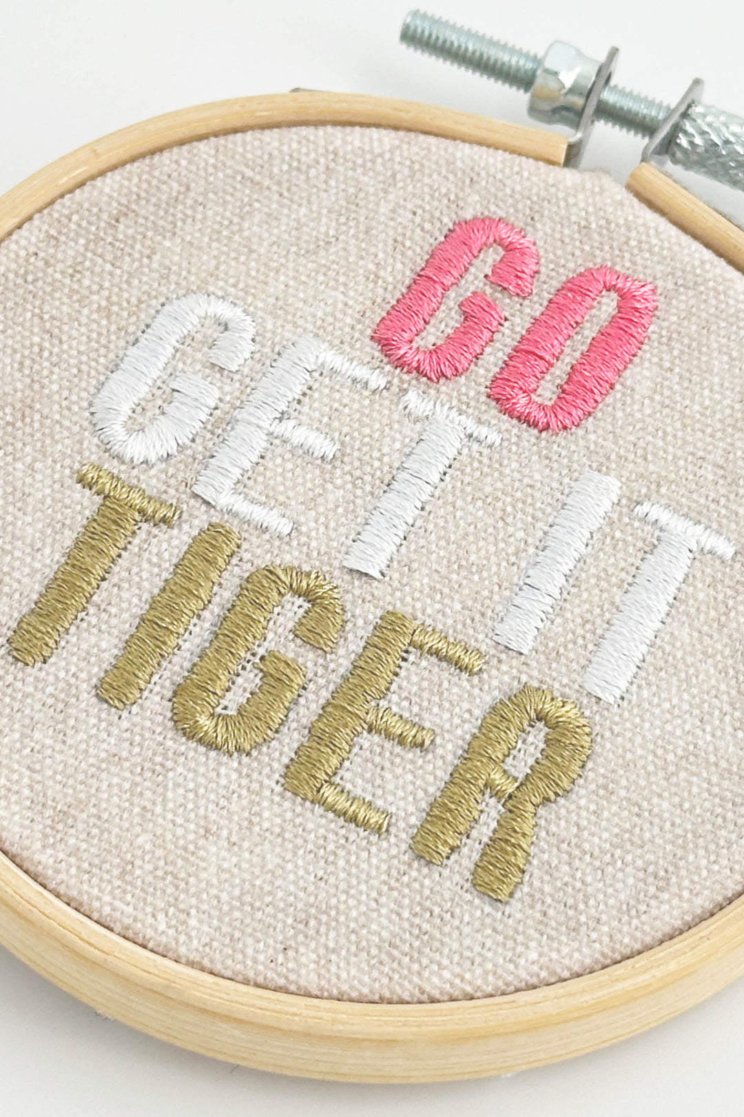 Embroidery hoop with 'Go Get It Tiger' text in pink, white, and gold on beige fabric.
