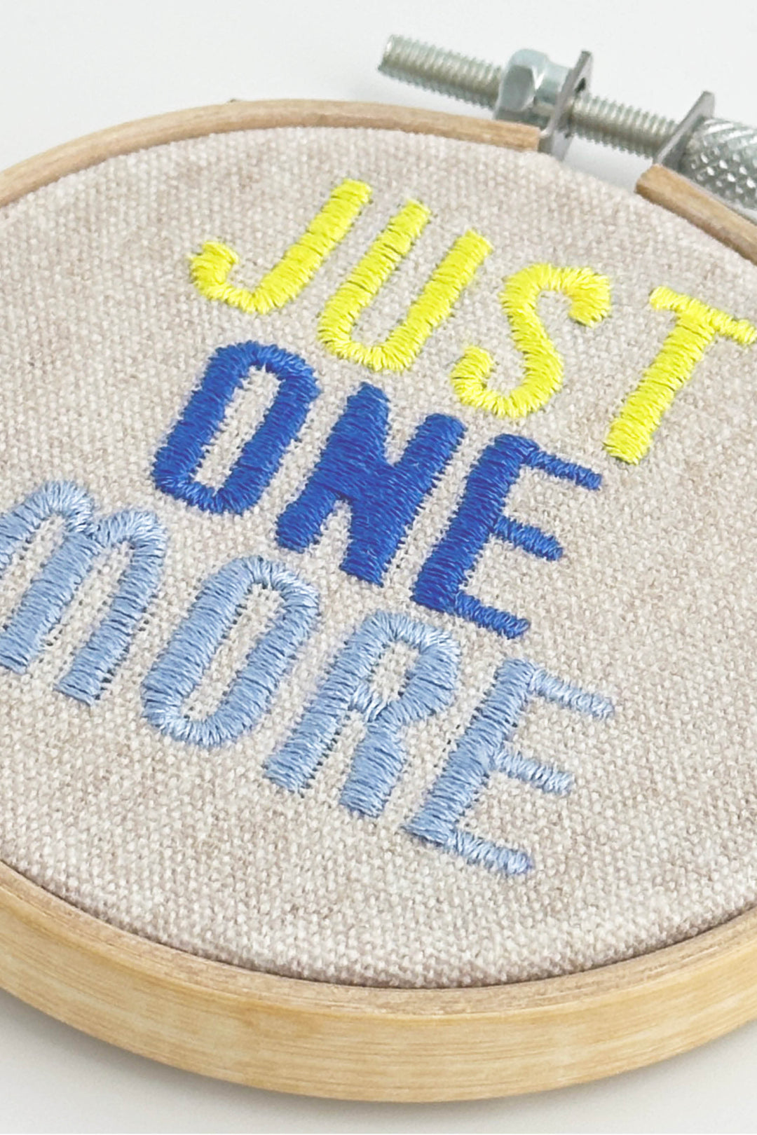 Embroidery hoop with 'Just One More' text in yellow, blue, and light blue on a white background