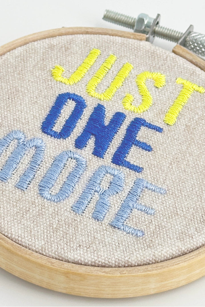 Embroidery hoop with 'Just One More' text in yellow, blue, and light blue on a white background