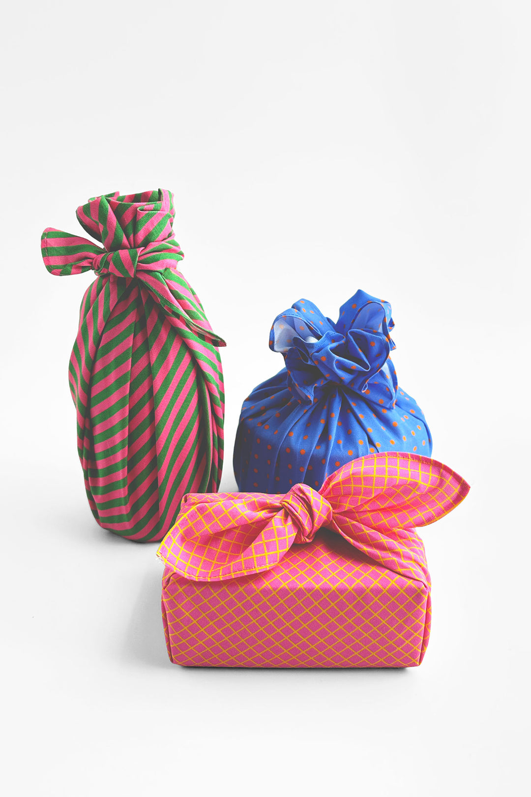 Three wrapped gift packages tied with bows on a clean white background: a pink and green striped cloth-wrapped package, a blue cloth-wrapped package, and a pink patterned gift box wrapped in matching pink ribbon.