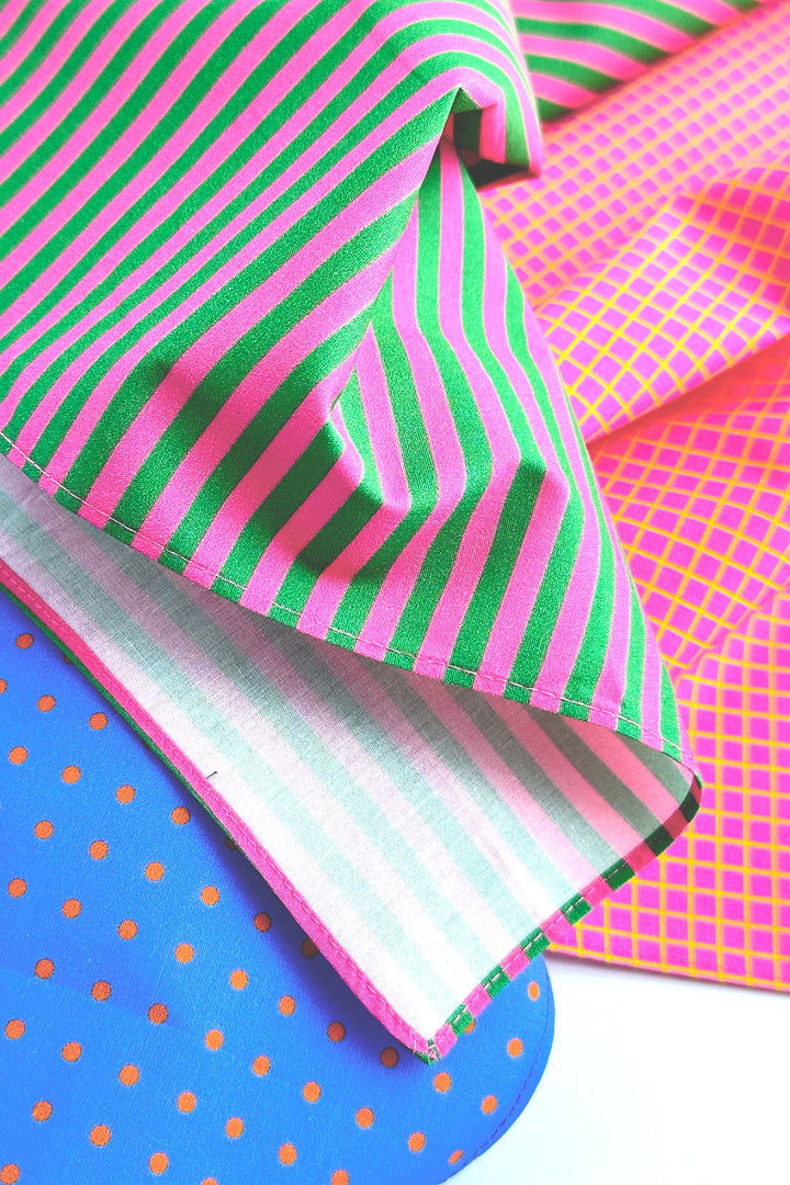 Three vibrant fabric samples layered diagonally: a pink and green striped fabric, a hot pink and yellow grid patterned fabric, and a blue fabric with orange polka dots.