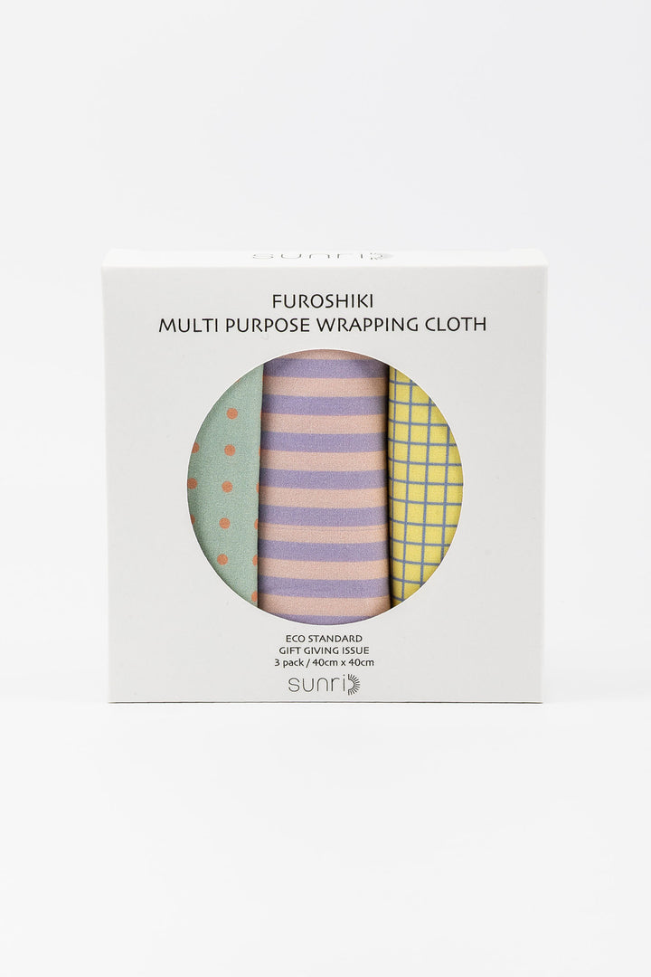 A boxed set of three folded wrapping cloths visible through a circular window on a white box. The cloths show pastel patterns: mint green with orange dots, lavender and pink stripes, and yellow grid.