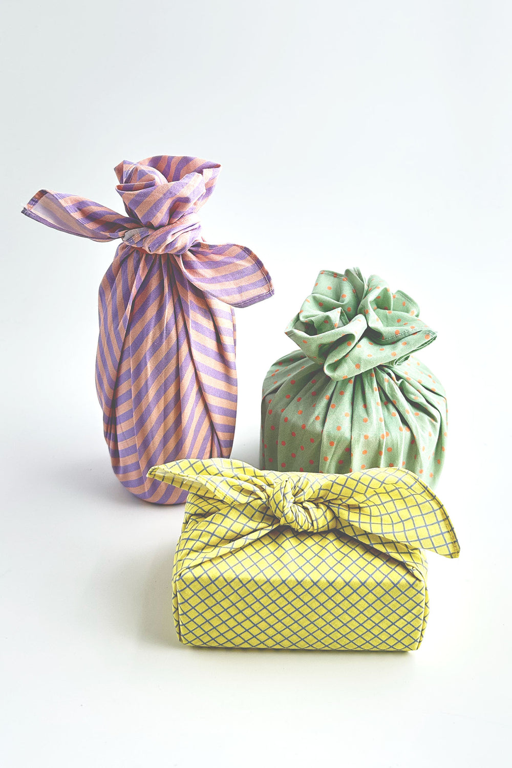Three fabric-wrapped packages tied with bows, arranged on a clean white background. The front package is yellow with a blue grid pattern, the left package is lavender and pink striped, and the right package is light green with small pink dots.