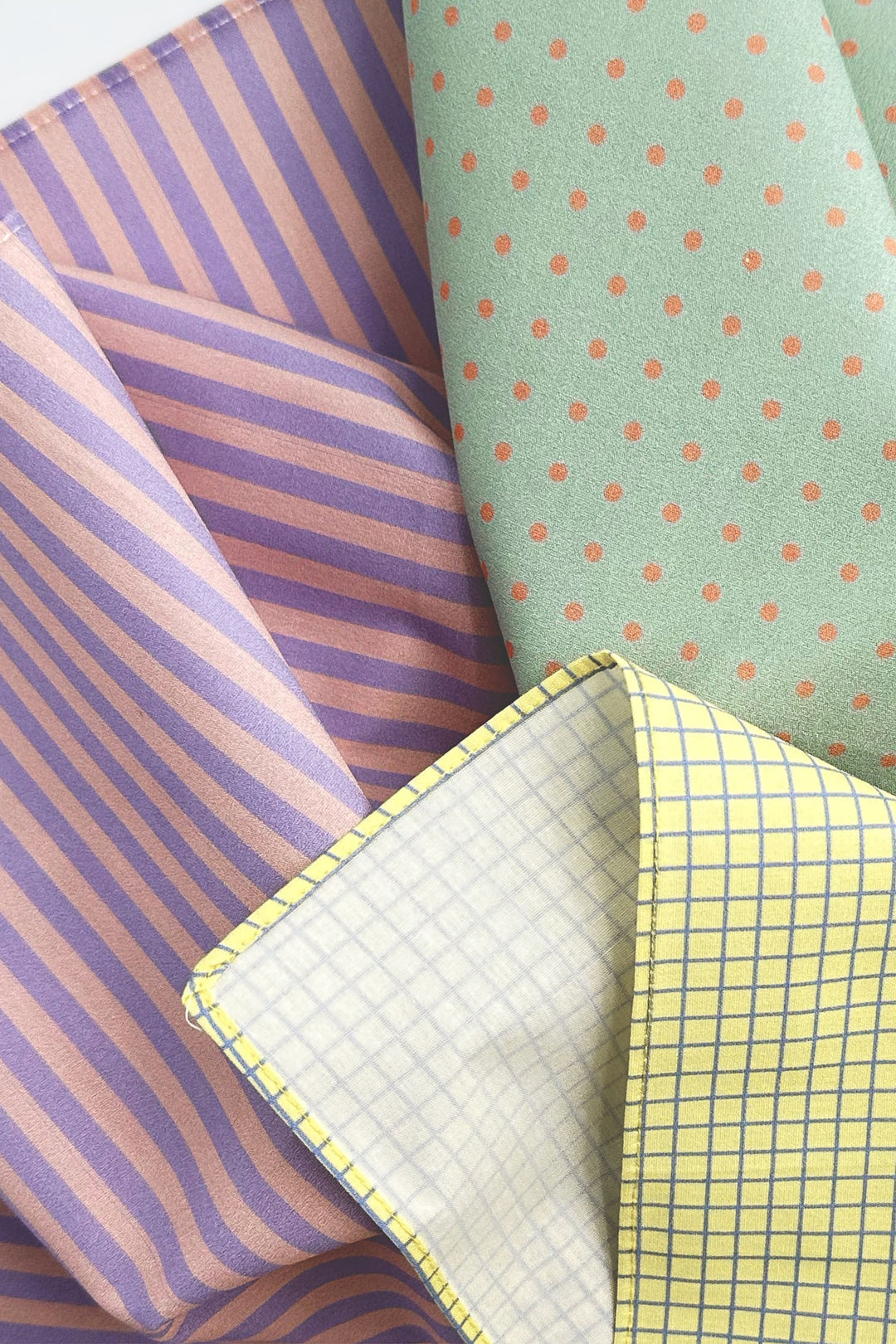 Four folded fabrics with different patterns: purple and pink stripes, mint green with orange polka dots, yellow and blue gingham, and a white fabric with a yellow grid pattern.