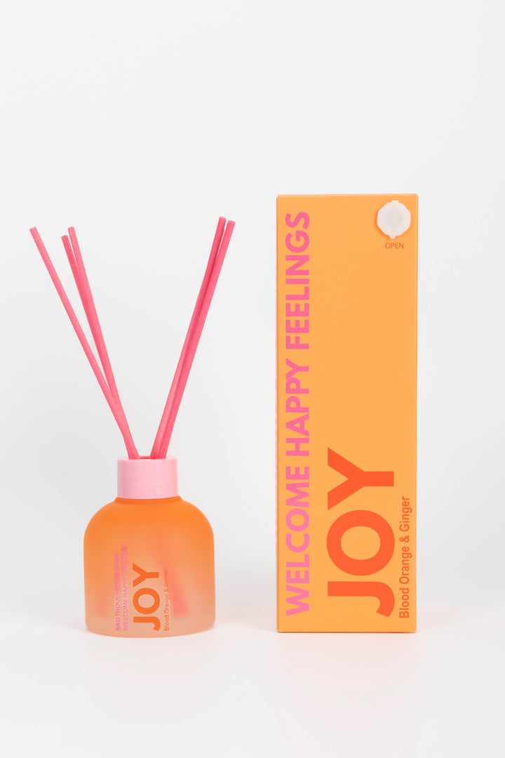 Reed diffuser with pink sticks and an orange bottle labeled 'JOY' next to its packaging on a white background.