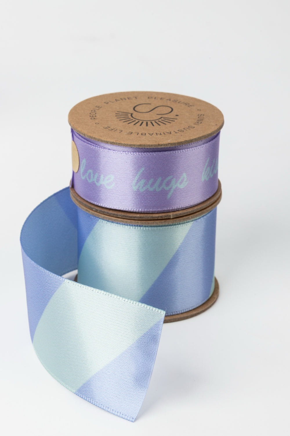 two satin ribbon rolls stacked with lilac upper roll and blue green diagonal lower roll partially unrolled
