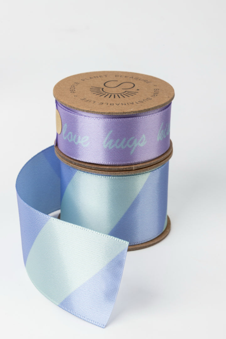 two satin ribbon rolls stacked with lilac upper roll and blue green diagonal lower roll partially unrolled
