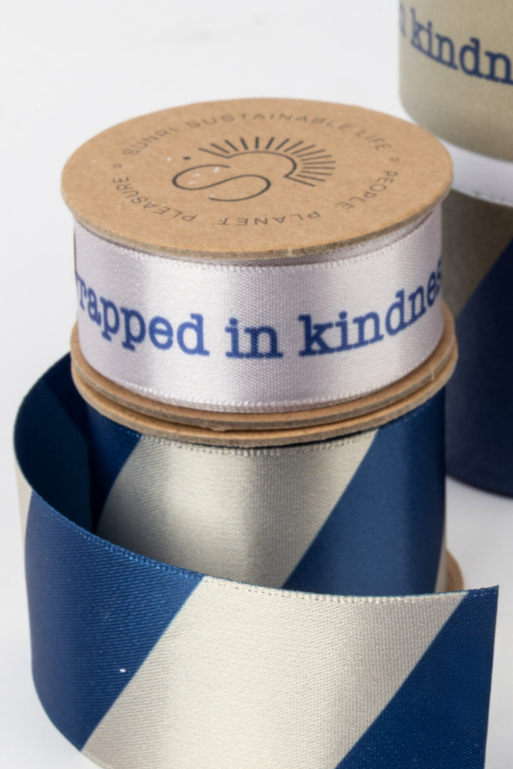 Spool of ribbon with 'wrapped in kindness' text and a branded lid.